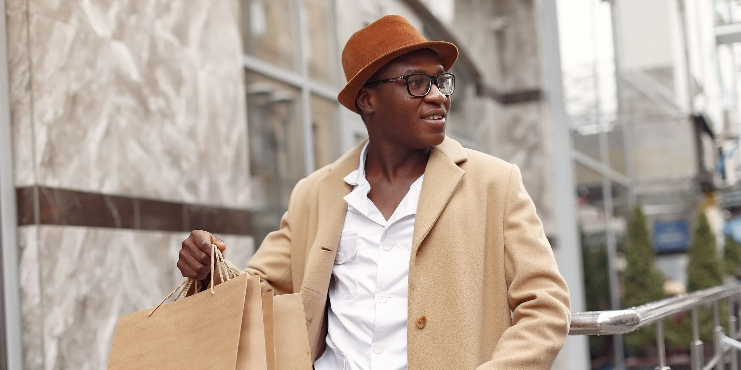 A smiling shopper in a camel coat and hat carrying kraft paper shopping bags down a city street.