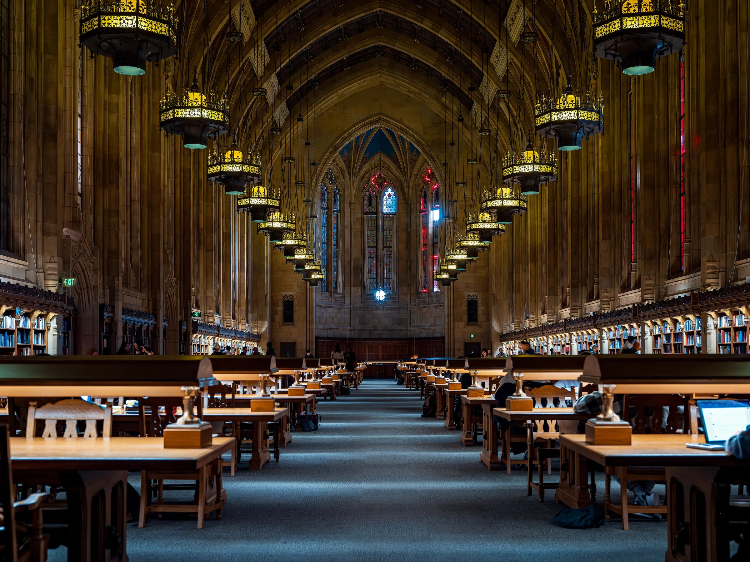 A long university reading room with rows of wooden desks, lamps and tall arched windows.